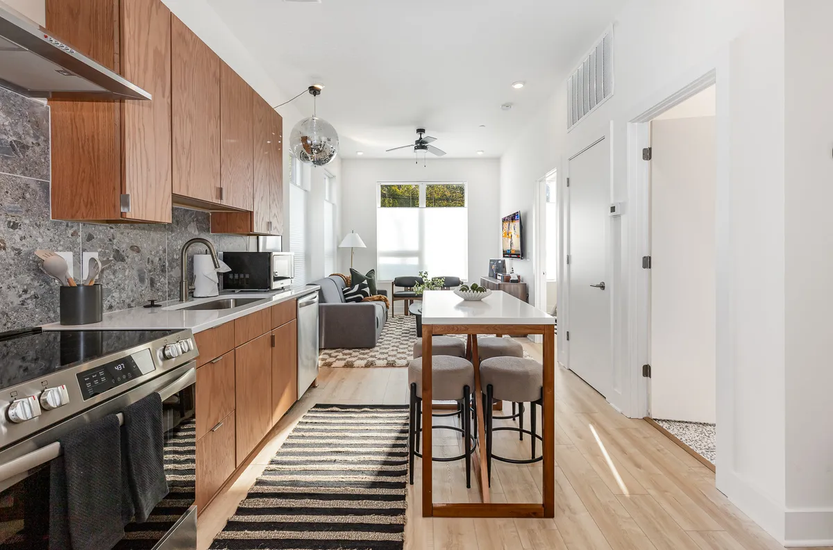 A contemporary, sunlit living space featuring a cozy sofa, a well-placed TV area, and a generous kitchen island with seating. Ample natural light streams in through the window, illuminating the seating and dining zones and giving the room a welcoming, lived-in feel. The integration of the kitchen and living area emphasizes the open-concept design mentioned in the section.
