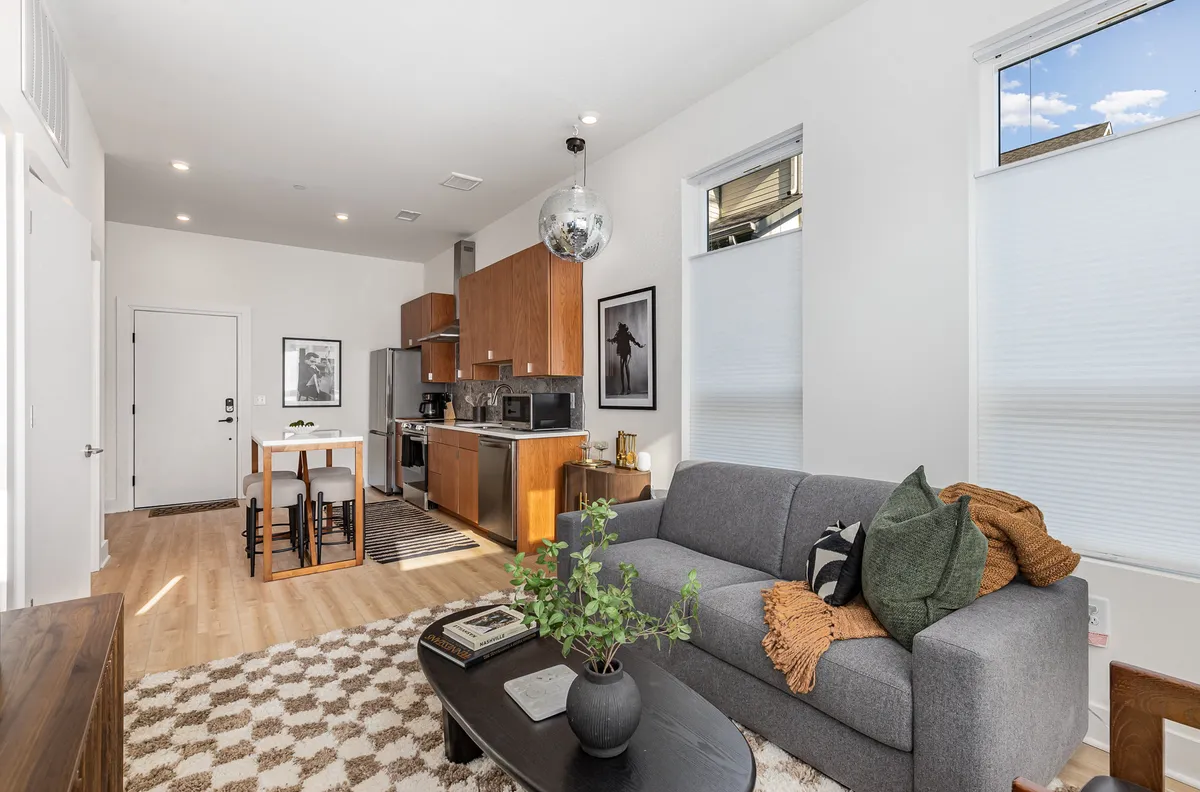 The image showcases a stylish open-concept living area seamlessly flowing into a modern kitchen. The scene features a plush sofa and coffee table in the foreground, a prominent kitchen island, striking wall artwork, and stainless steel appliances in the background. The space is bathed in natural light, exuding a warm and inviting atmosphere ideal for socializing or relaxing after a day out.