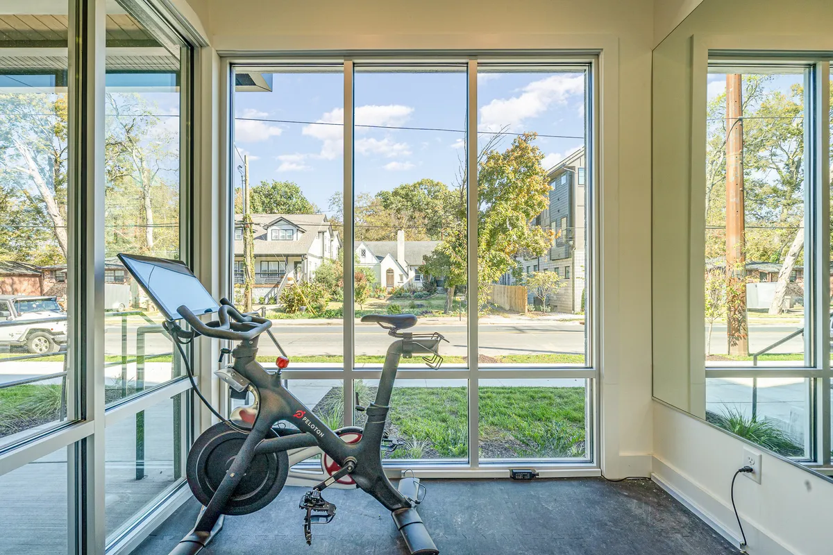 A modern fitness room flooded with natural light. The focal point is a high-end exercise bike positioned near expansive windows, with a wall mirror reflecting the spaciousness of the room. The setup includes a Peloton bike and Echelon fitness mirror, offering a motivating and high-tech environment for workouts.