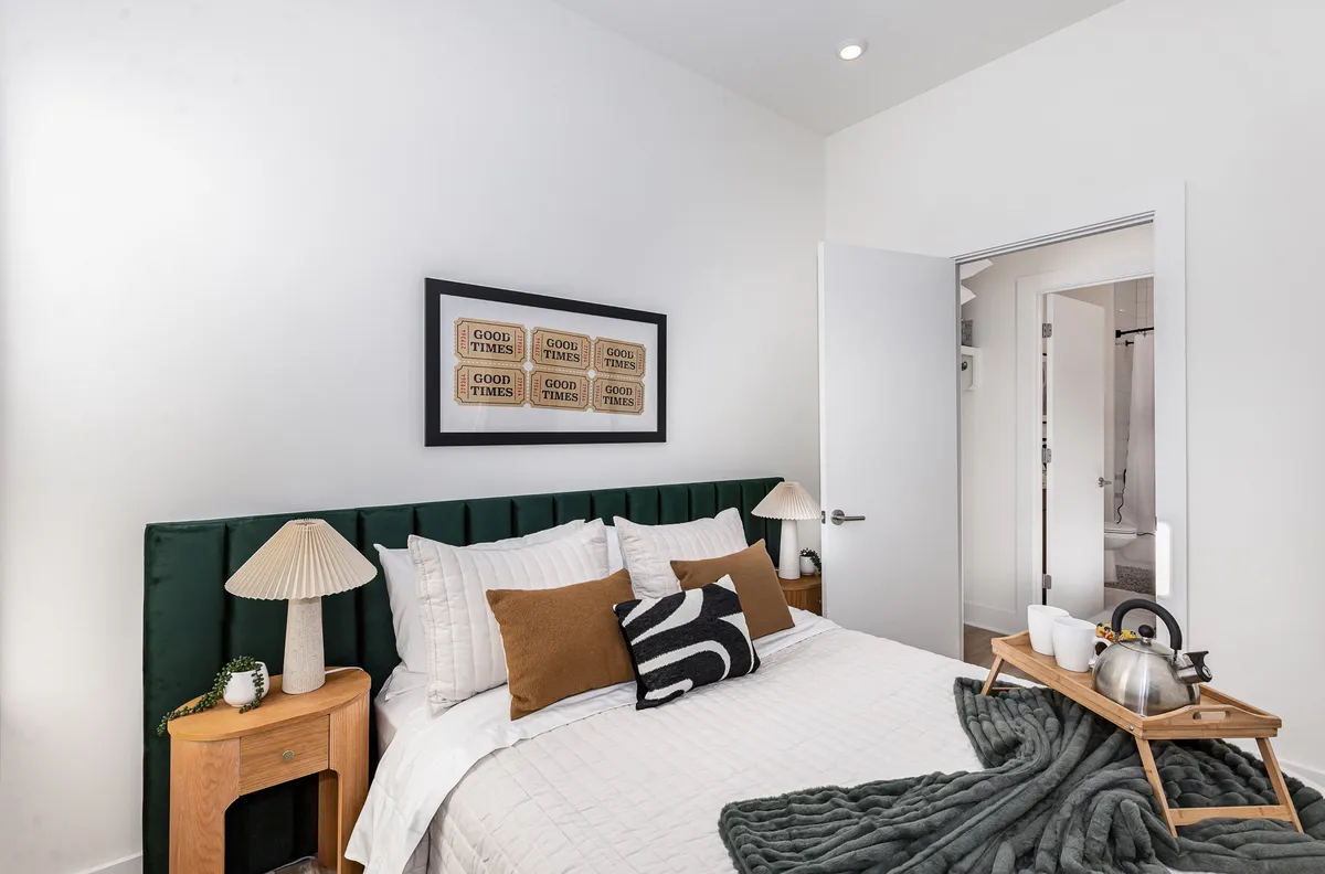 A close-up view of a comfortable bed with a green headboard, layered with decorative pillows and a nightstand with a glowing lamp. The setting is intimate and warm, suggesting a peaceful bedtime routine.