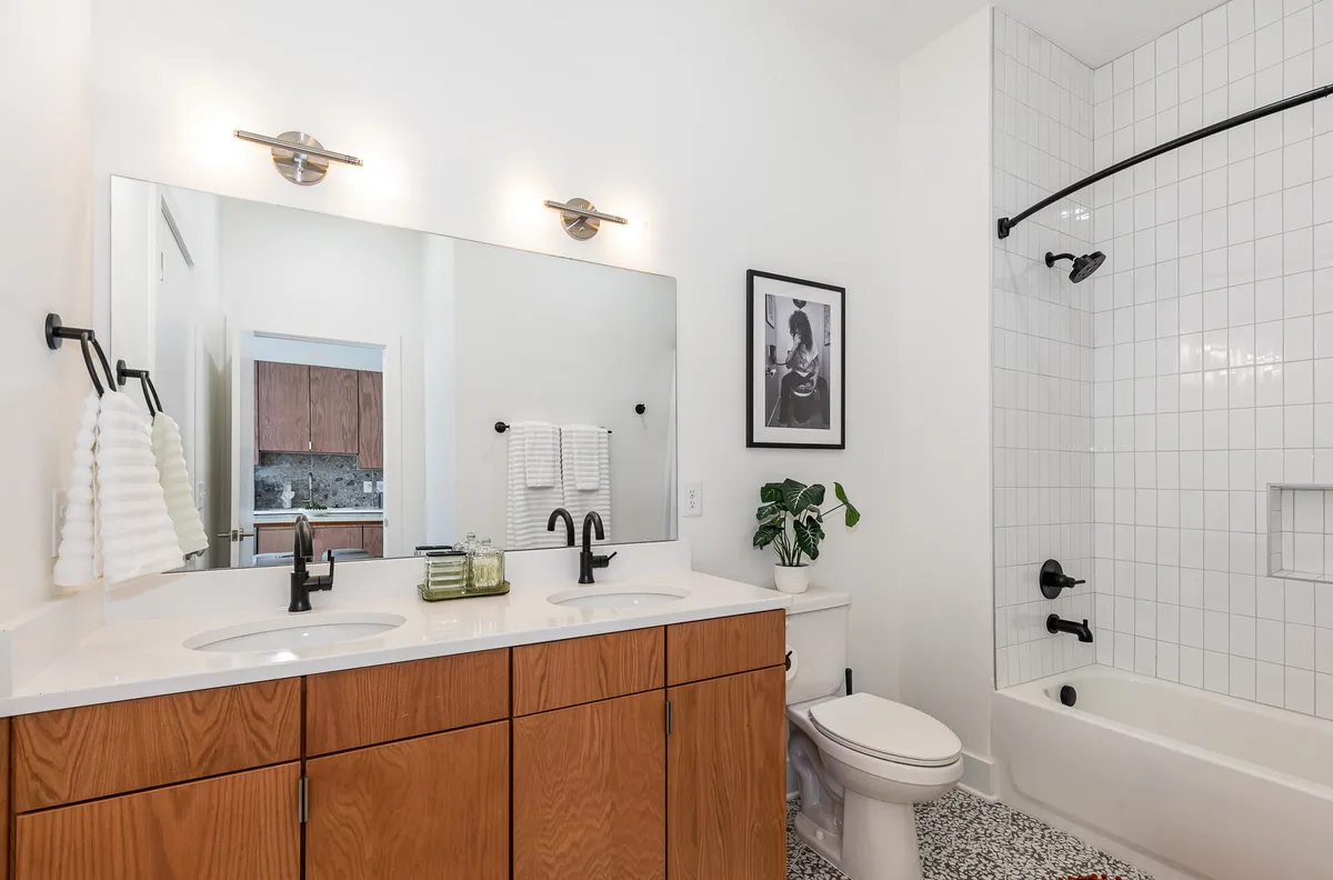 A tranquil, modern bathroom with a double sink vanity, large mirror, tasteful artwork, and a green plant. The fixtures and cleanliness evoke a spa-like ambiance, ideal for relaxing and refreshing after your journey.