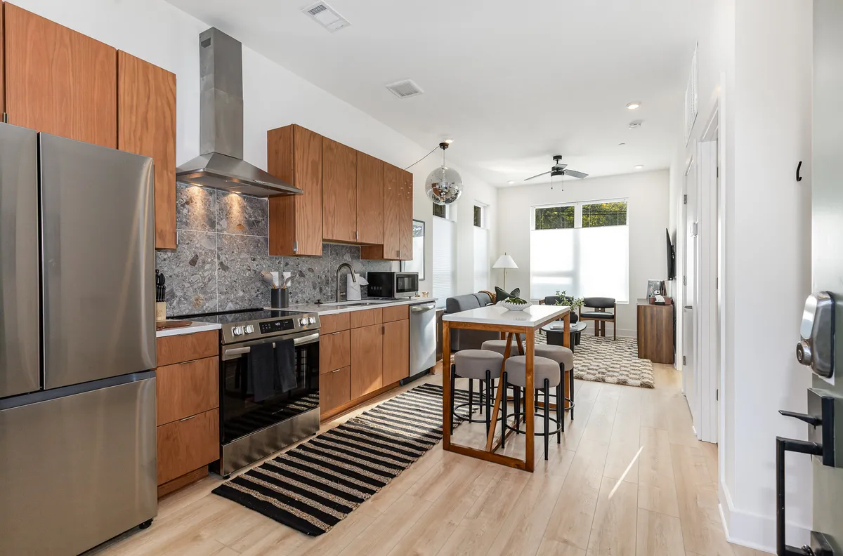This inviting interior features a modern kitchen with stainless steel appliances, warm wooden cabinetry, a cozy dining table set atop a striped rug, and ample space for cooking, dining, and socializing with loved ones.
