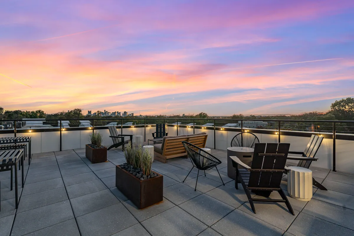 A serene exterior scene of a rooftop with outdoor seating, lush planters, and a backdrop of the city skyline at sunset. The colorful sky and comfortable arrangement suggest a tranquil spot for evening gatherings or a peaceful morning coffee.
