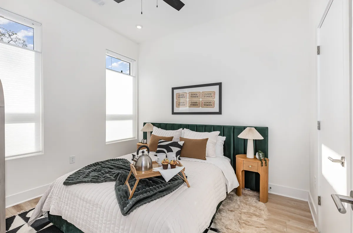 A serene bedroom featuring a plush bed with a green headboard and luxurious linens. Decorative wall art adds personality, while a tray with a kettle and breakfast items sits atop the bed. Soft natural light streams in from expansive windows, creating a peaceful and rejuvenating retreat.